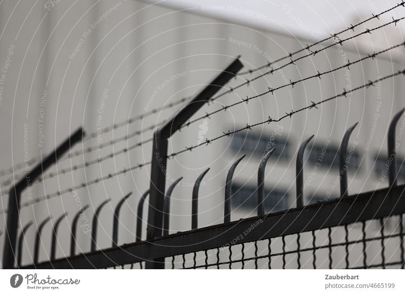 Zaun mit Stacheldraht, dahinter graue Fassade Grenze Stäbe Metall Barriere Schutz Sicherheit bedrohlich Stacheldrahtzaun gefangen gefährlich Angst Freiheit