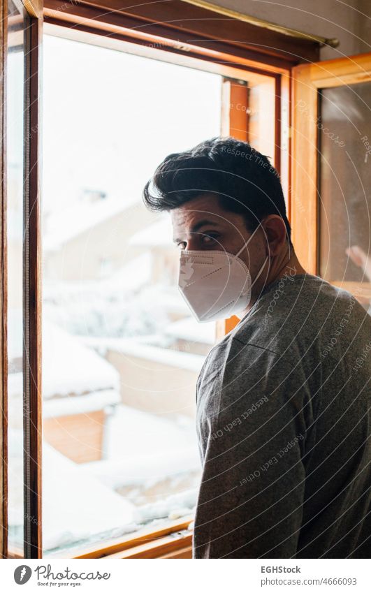 Junger Mann mit Maske in Quarantäne, der durch das Fenster seines Hauses den Schneefall betrachtet allein Angst begrenzt Einsperrung Kontemplation Korona