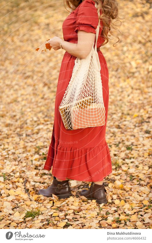 Schöne Frau im Herbst Park trägt ein rotes terracota Farbe Kleid, stehend auf Herbstblätter mit Öko-Netz Tasche mit Büchern. Junge Millennial-Frau mit langen Haaren in stilvollen Herbst-Outfit, Blick in die Kamera. Herbst weiblichen Lebensstil, Inspiration.