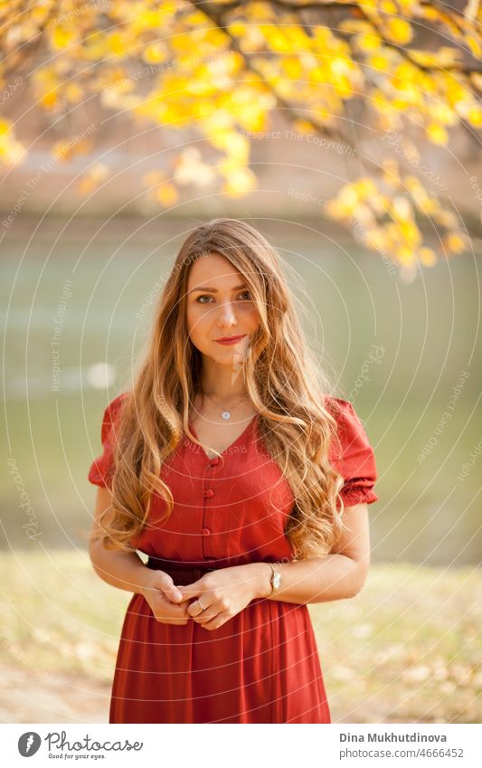 Schöne Frau im Herbst Park trägt eine rote terracota Farbe Kleid, stehend im Herbst Park in der Nähe von See mit gelben Laub hinter ihr. Junge Millennial Frau mit langen Haaren in stilvollen Herbst-Outfit, Blick in die Kamera. Herbst weiblichen Lebensstil, Inspiration.