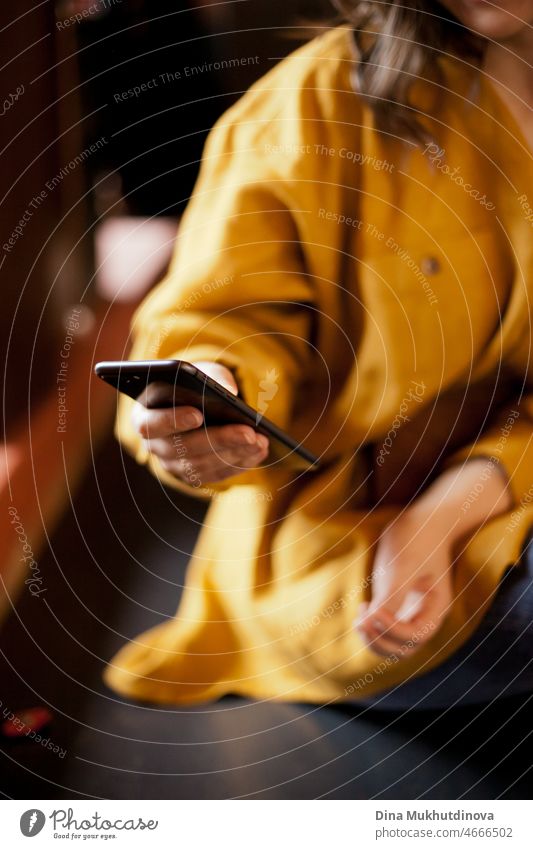 Schwarzes Mobiltelefon in weiblicher Hand, Nahaufnahme. Unerkennbare Frau in gelber Kleidung sitzt in einem Café und schreibt eine SMS auf dem Handy. Echte Menschen benutzen Mobiltelefone an öffentlichen Orten. Millennial und Generation Z digitalen Lebensstil.