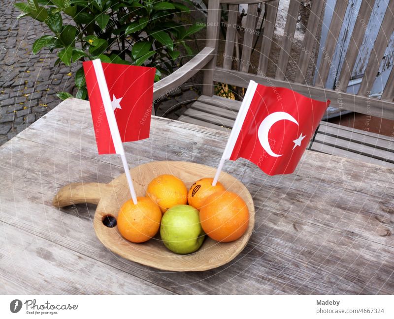 Frische Orangen und grüner Apfel mit rotentürkischen Fähnchen in einer dekorativen Holzschale auf einem rustikalen Holztisch mit Holzbank in den Gassen der Altstadt von Alacati bei Izmir in der Türkei