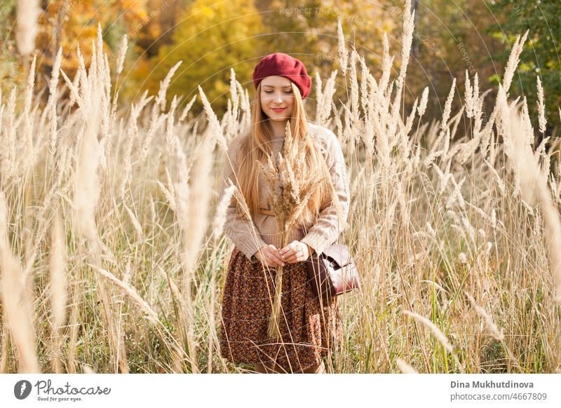 Schöne Frau im Herbst Park trägt eine weinrote Baskenmütze, beige Pullover und braunen Rock, stehend in trockenen Weizen wildes Feld. Junge Millennial-Frau mit langen Haaren in stilvollen Herbst-Outfit, lächelnd und Blick in die Kamera. Herbst weiblichen Lebensstil, Inspiration.