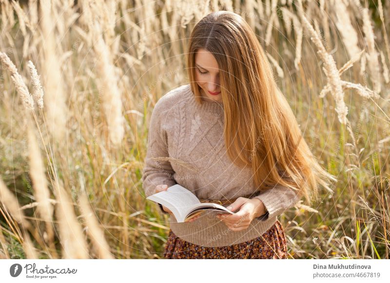 Schöne Frau beim Lesen eines Buches trägt einen beigen Pullover und braunen Rock, stehend in trockenen Weizen wildes Feld. Junge Millennial-Frau mit langen Haaren in stilvollen Herbst-Outfit, lächelnd und Blick in die Kamera. Herbst weiblichen Lebensstil, Inspiration.