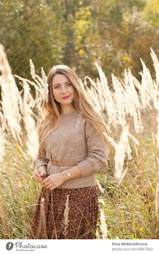 Schöne Frau im Herbst Park trägt einen beigen Pullover und braunen Rock, stehend in trockenen Weizen wildes Feld. Junge Millennial-Frau mit langen Haaren in stilvollen Herbst-Outfit, lächelnd und Blick in die Kamera. Herbst weiblichen Lebensstil, Inspiration.