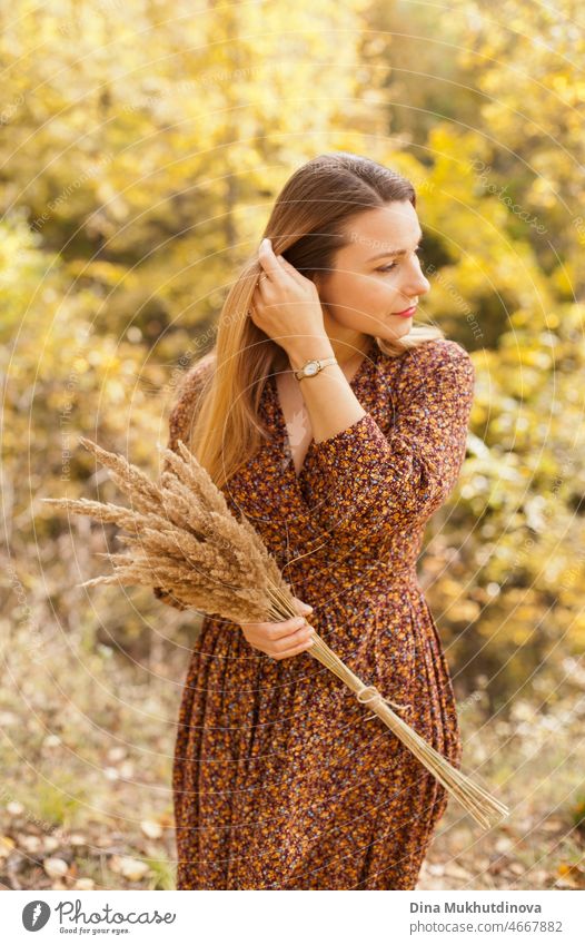 Schöne Frau im Herbst Park trägt ein braunes Kleid, hält einen trockenen Weizen Bouquet. Junge Millennial-Frau mit langen Haaren in stilvollem Herbst-Outfit, lächelnd und Blick zur Seite. Herbst Lebensstil, Inspiration und Weiblichkeit.