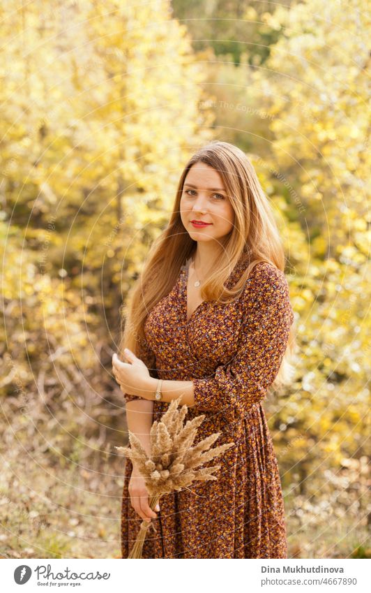 Schöne Frau im Herbst Park trägt ein braunes Kleid, hält einen trockenen Weizen Bouquet. Junge Millennial-Frau mit langen Haaren in stilvollen Herbst-Outfit, lächelnd und Blick in die Kamera.