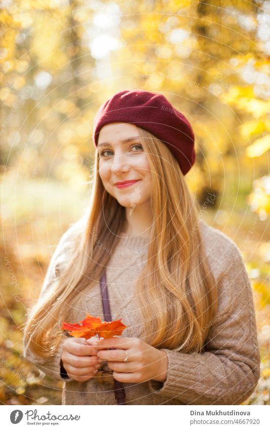 Schöne Frau im herbstlichen Park, die eine weinrote Baskenmütze trägt und ein rotes Ahornblatt hält. Junge Frau mit langen Haaren in stilvollem Herbst-Outfit, lächelnd und in die Kamera blickend.