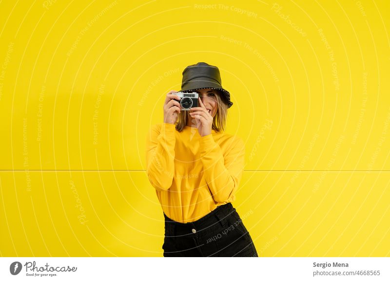 Nahaufnahme einer jungen kaukasischen Frau mit schwarzem Hut, auf gelbem Hintergrund, die eine alte Fotokamera hält Porträt schön Menschen Kaukasier Person