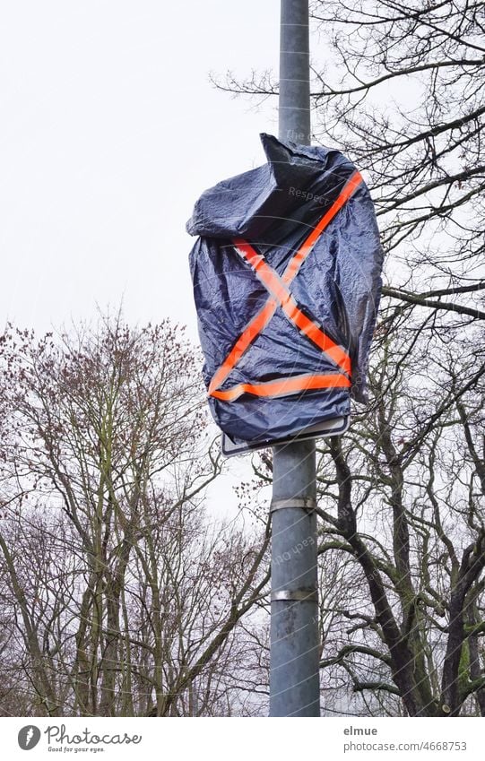 Verkehrsschild und Zusatzzeichen, verhüllt mit einer blauen Plastiktüte und orangefarbenen Klebestreifen vor kahlen Bäumen / Straßenverkehr / Baumaßnahme