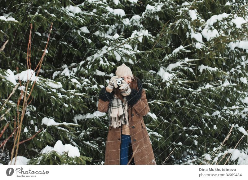 Schöne Frau mit langen braunen Haaren, die Fotos auf Film Retro-Kamera zu Fuß im Winter Park mit Tannen während Schneefall. Wintermode und stilvolles Outfit. Echte Menschen, die Spaß im Winter, genießen die frische Luft in der Natur mit verschneiten Fichten.
