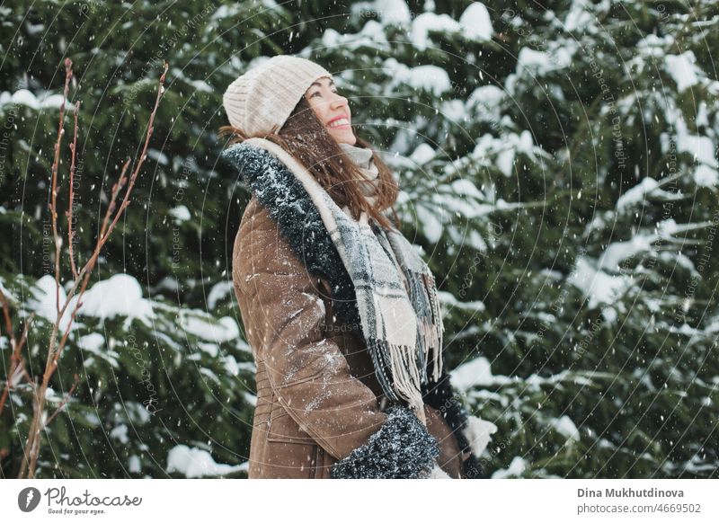Schöne Frau mit langen braunen Haaren zu Fuß im Winter Wald oder Park mit Tannen bei Schneefall. Wintermode und stilvolles Outfit. Echte Menschen, die Spaß im Winter, genießen die frische Luft in der Natur mit verschneiten Fichten.