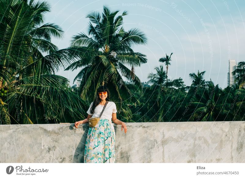 Lächelnde Frau mit Sonnenbrille lehnt an einer Mauer vor Palmen. Gute Laune im Urlaub. lächelnd gute Laune Sommer Fröhlichkeit Tropen Urlaubsstimmung positiv