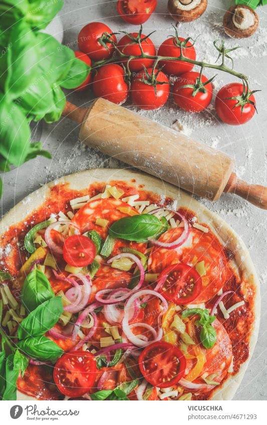 Nahaufnahme einer hausgemachten Pizza mit Tomaten, Salami, Zwiebeln und Basilikum am Küchentisch Italienische Küche selbstgemacht Tisch Mehl hölzern Nudelholz