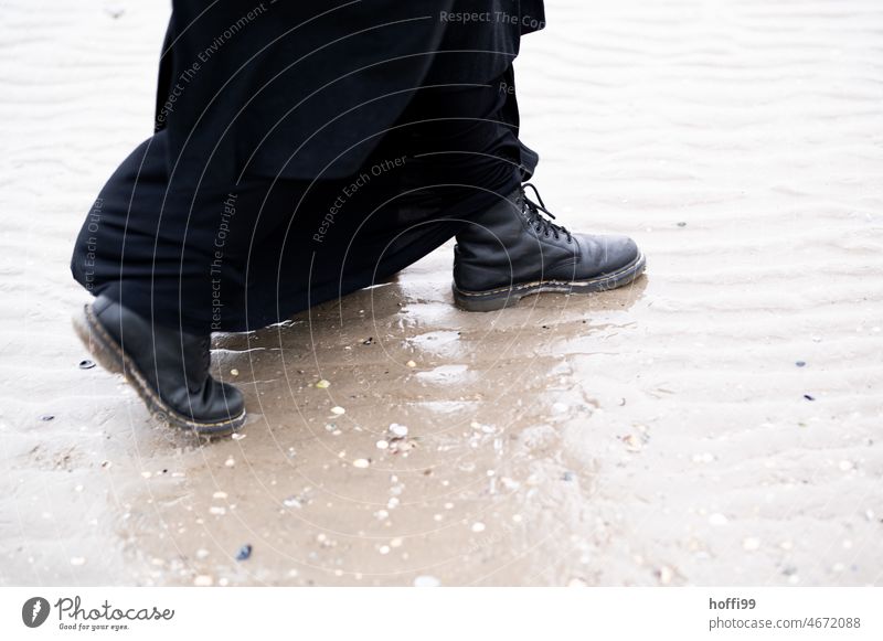 Schwarze Stiefel mit langem Mantel schreiten bei Ebbe durchs Watt Schritte Wattenmeer Wasser Pfütze schwarz schwarze Stiefel schwarzer Mantel Minimalismus gehen
