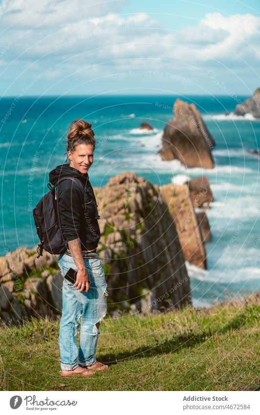 Lächelnde Frau steht an der Küste mit blauem Meerwasser Reisender MEER Hügel Klippe Fernweh Freiheit positiv erkunden Natur Smartphone Ausflug Tourismus reisen