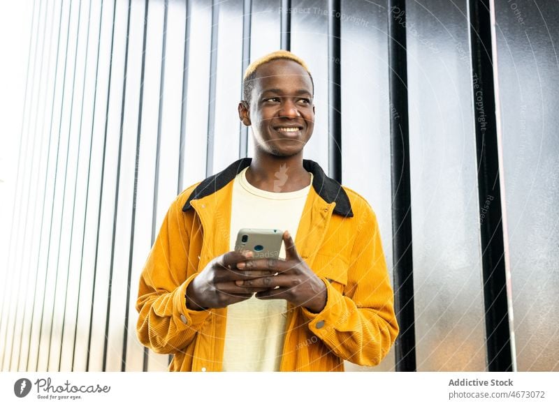 Fröhlicher schwarzer Mann beim Surfen auf dem Smartphone Browsen online Textnachricht Stil Wand Talkrunde digital Straße Vorschein Mode gefärbtes Haar modern
