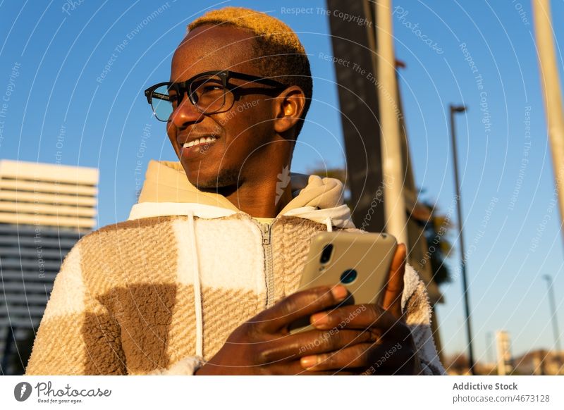 Lächelnder schwarzer Mann surft mit Smartphone in der Stadt Browsen online Textnachricht Talkrunde digital Straße Großstadt Gebäude Mode gefärbtes Haar modern