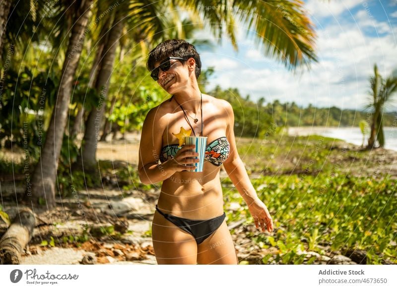 Positive Frau im Bikini mit Getränk an der Küste Badebekleidung Erfrischung Ufer Erholung Natur MEER tropisch Baum ruhen Tourist Zeitvertreib Ausflug Wasser