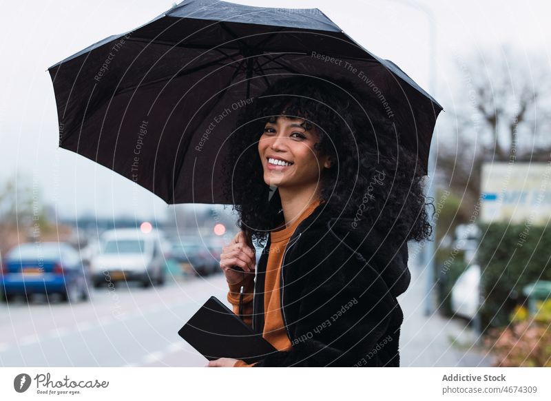 Lächelnde schwarze Frau mit Regenschirm auf der Straße Großstadt Straßenrand urban Vorschein Tablette Porträt afroamerikanische Frau lässig jung Glück positiv