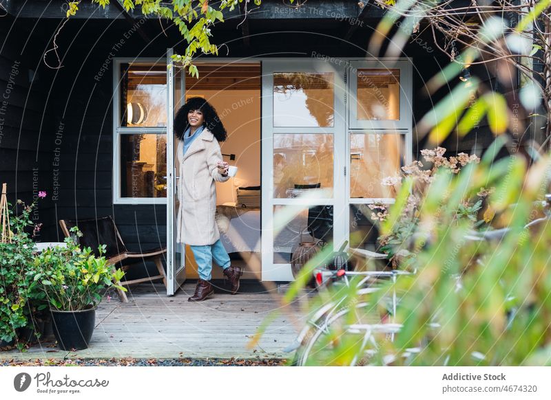 Glückliche schwarze Frau steht in der Tür eines Hauses heimwärts hinausgehend warme Kleidung Mantel Oberbekleidung Türöffnung urban Kaffee Tasse Getränk Tee