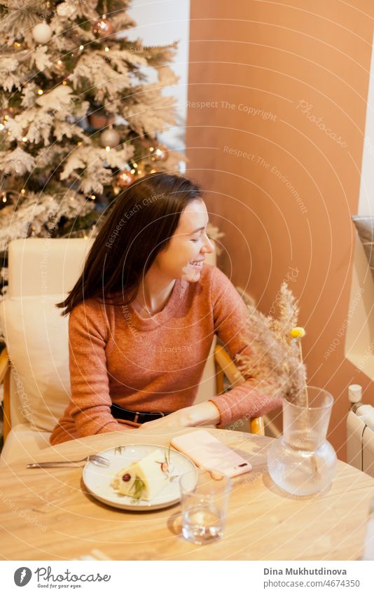Junge Frau sitzt an einem Tisch im Café mit Weihnachtsbaum Lichter hinter ihr, essen einen Kuchen am Valentinstag. Junge Frau im Cafe offener Lebensstil schön
