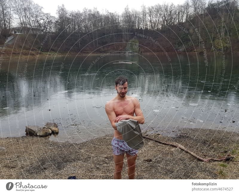 junger Mann mit nacktem Oberkörper an einem vereisten Badesee Eisbaden Winter eisschwimmen immunsystem frieren kalt ausziehen anziehen Badehose abhärten