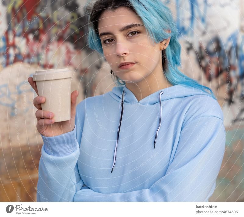 Blauhaariges Teenager-Mädchen in hellblauem Oversize-Kapuzenpulli in der Nähe einer Graffiti-Wand mit Kaffee zum Mitnehmen hell-blau Attrappe Übergröße Tasse
