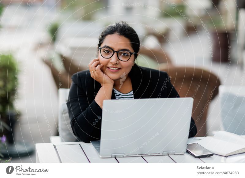 Eine junge und selbstbewusste indische Asiatin lächelt, während sie an einem Sommertag in einem schicken Straßencafé an ihrem Laptop arbeitet. Mädchen Inder
