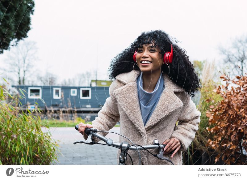 Fröhliche schwarze Frau mit Kopfhörern und Fahrrad benutzend Musik zuhören Kälte sich[Akk] entspannen Großstadt afroamerikanische Frau Straße Lächeln