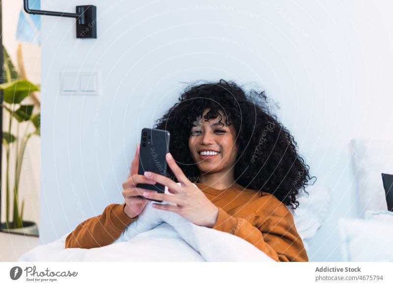 Glückliche schwarze Frau benutzt Smartphone im Bett benutzend wach Morgen Wochenende zu Hause sich[Akk] entspannen ruhen Afroamerikaner Mobile Telefon Browsen