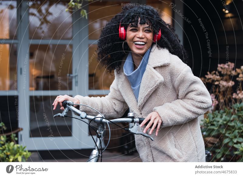 Fröhliche schwarze Frau mit Kopfhörern und Fahrrad benutzend Musik Lachen zuhören Kälte sich[Akk] entspannen Großstadt afroamerikanische Frau Straße Lächeln