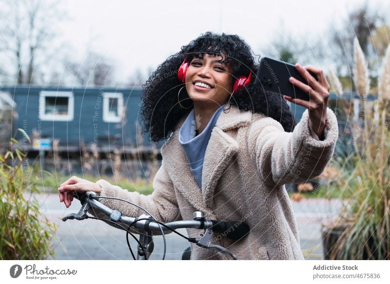 Vergnügte schwarze Frau mit Smartphone auf dem Fahrrad Kopfhörer benutzend Musik unter Selfie zuhören Mitfahrgelegenheit Kälte afroamerikanische Frau