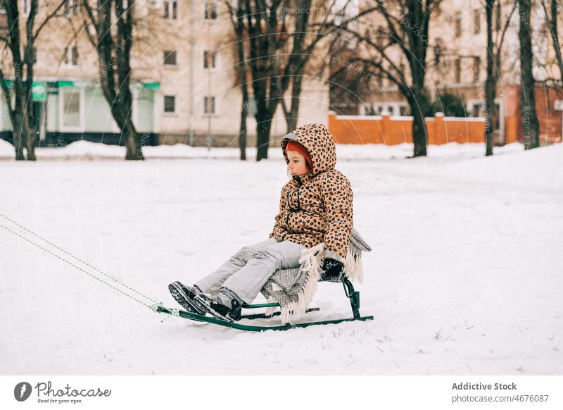 Kind auf Schlitten sitzend an einem Wintertag Kindheit Schnee Straße Großstadt Saison Frost Oberbekleidung kalt Wetter warme Kleidung Temperatur gefroren