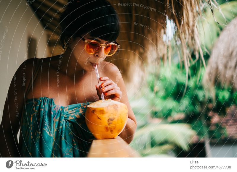 Lächelnde, in buntes Badetuch gewickelte, Frau mit Sonnenbrille trinkt aus einer King Kokosnuss mit Strohhalm in der Hand. Urlaub in den Tropen. Trinkkokosnuss