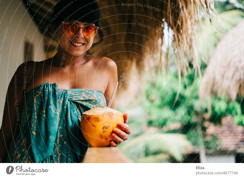 Lächelnde, buntes Badetuch gewickelte, Frau mit Sonnenbrille hält eine King Kokosnuss mit Strohhalm in der Hand. Urlaub in den Tropen. Trinkkokosnuss Sommer
