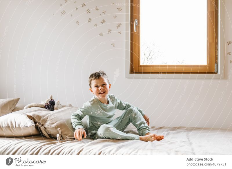 Fröhlicher Junge auf dem Bett sitzend Kind Fenster Schlafzimmer Pyjama Kindheit heimisch Morgen Spaß haben lustig Appartement bezaubernd flach niedlich