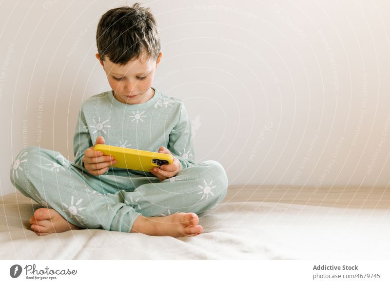 Junge im Pyjama, der auf seinem Smartphone spielt Kind Kindheit Bett Schlafzimmer Videospiel spielen Zeitvertreib Morgen heimisch Appartement bezaubernd Barfuß