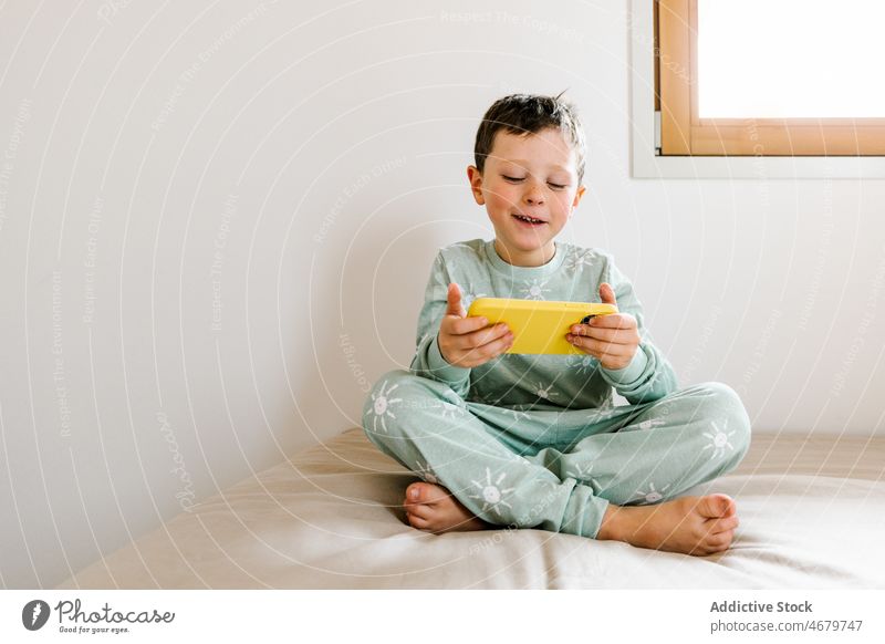 Fröhlicher Junge im Pyjama, der mit seinem Smartphone spielt Kind Kindheit Bett Schlafzimmer Videospiel spielen Zeitvertreib Morgen heimisch Appartement