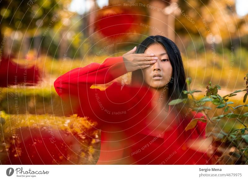 Porträt einer ostasiatischen Frau, die einen roten Anzug trägt und ihr Auge mit einer Hand in einem Garten bedeckt elegant posierend Spanien Blumen Park