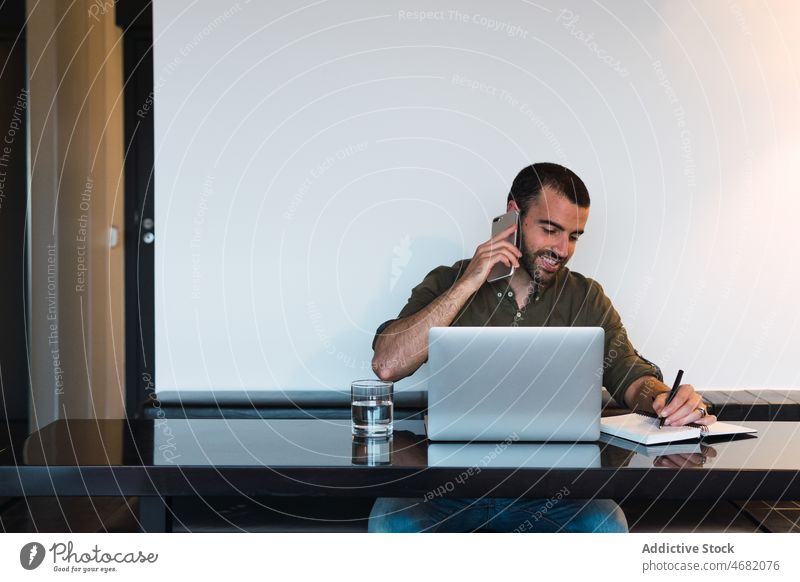 Fröhlicher Mann, der in der Nähe eines Laptops mit seinem Smartphone spricht Gespräch Telefonanruf Arbeit Arbeitsplatz Talkrunde Kommunizieren diskutieren