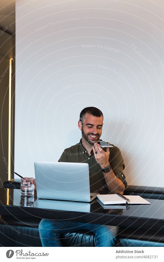 Positiv eingestellter Mann, der in der Nähe seines Laptops eine Sprachnachricht aufzeichnet Smartphone Aufzeichnen Stimme Audio Nachricht Arbeit Arbeitsplatz