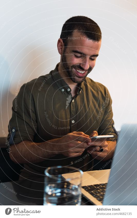 Glücklicher Mann, der in der Nähe seines Laptops auf seinem Smartphone surft Browsen online Internet Lächeln Arbeit Arbeitsplatz modern froh Freude