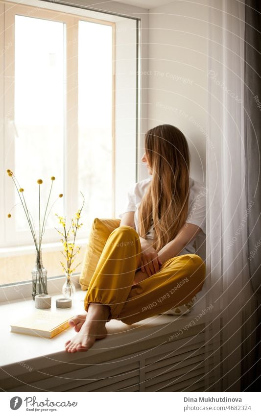 Junge schöne Frau in weißem T-Shirt und gelber Jogginghose liest ein Buch zu Hause, sitzt auf der Fensterbank, lächelnd. Gemütlich entspannt lässigen Lebensstil Ganzkörper-Porträt. Literatur und belletristische Bücher.  Gemütliches Zimmer zu Hause.