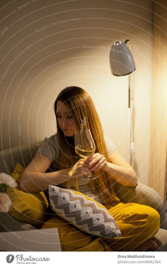 Junge attraktive Frau sitzt zu Hause in coxy Stuhl trinken Weißwein. Verwöhnen Sie sich nach einem harten Arbeitstag. Verkostung von Wein. Gelbe Farbe im Haus Interieur. Casual alltäglichen Lebensstil Millennials.
