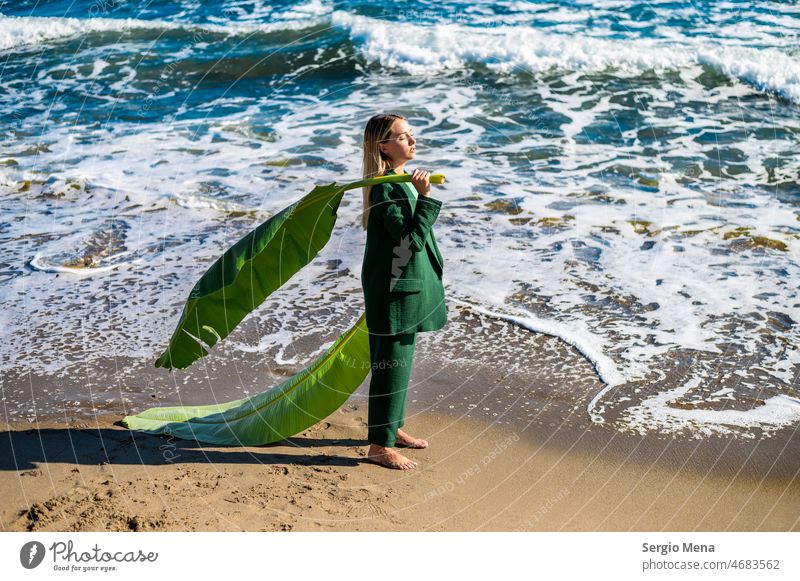 Künstlerische Fotografie einer eleganten kaukasischen Frau am Strand mit einem großen grünen Blatt Nahaufnahme Mädchen blond hübsch schön attraktiv Stehen