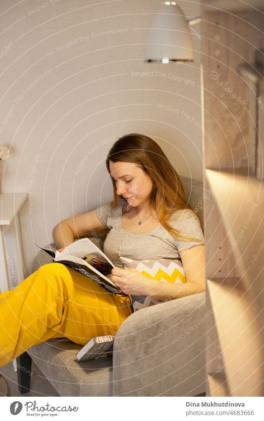 Junge schöne Frau trägt grau und gelb Jogginghose ein Buch zu Hause zu lesen, sitzen in gemütlichen Stuhl. Literatur und belletristische Bücher.  Gemütliches Zimmer zu Hause.