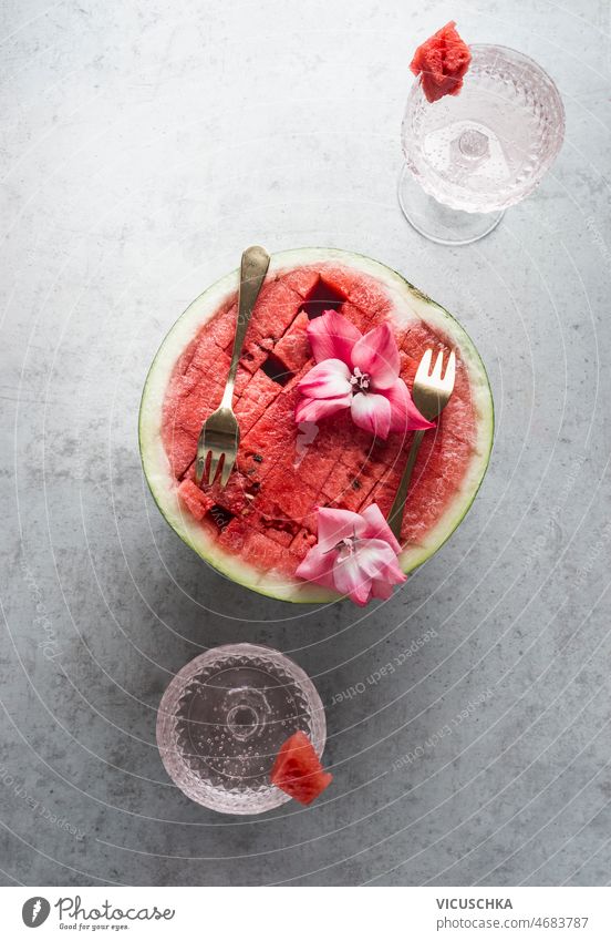 Halbierte Wassermelone mit goldenen Gabeln und Blumen am grauen Betonküchentisch mit Sektgläsern halbiert Küche Tisch Champagne Brille Sommer Lebensmittel