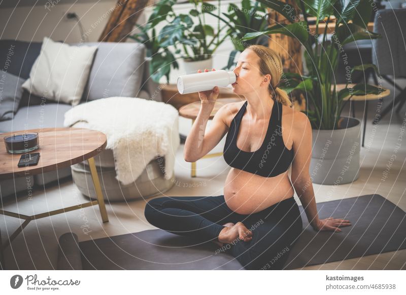 Junge glückliche und fröhliche schöne schwangere Frau, die eine Pause macht, hydratisiert und Wasser aus der Flasche trinkt, nachdem sie zu Hause ein Wellness-Trainingsprogramm absolviert hat