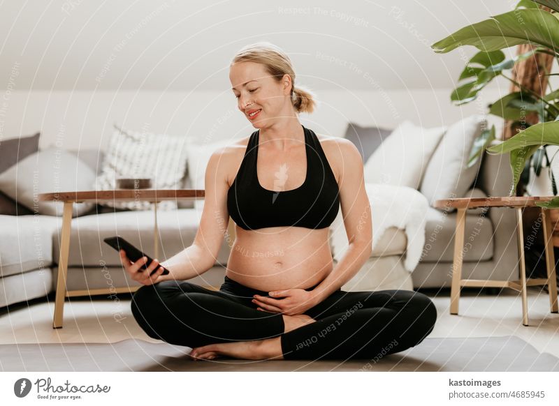 Schwangerschaft und technologiegestützte gesunde Lebensweise Konzept. Fröhlich glücklich schwangere Frau mit Smartphone-Anwendung während der Ausübung auf Yoga-Matte auf Wohnzimmerboden zu Hause.